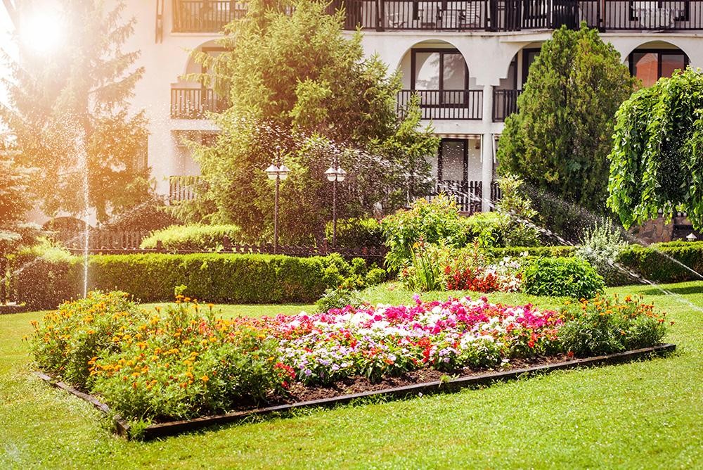 Well-kept garden with colorful flower beds, trimmed hedges, and a sunlit building in the background.